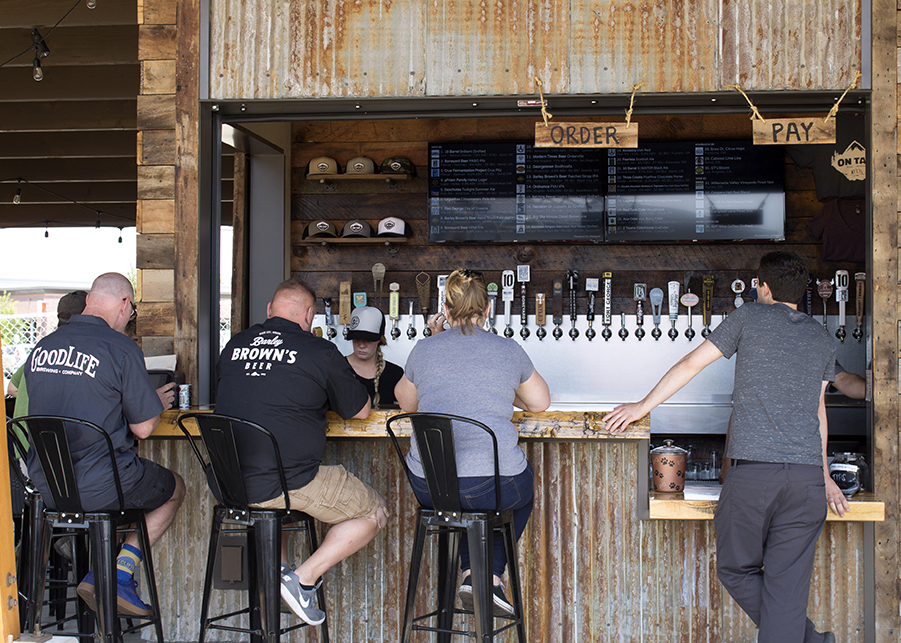 Several patrons sit at the bar constructed from exposed wooden planks and tin siding allowed to oxidize naturally